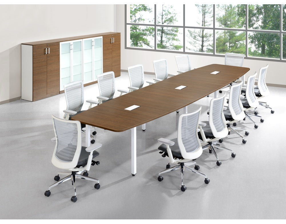 A modern conference room setup featuring a long rectangular wooden table with white metal legs, surrounded by white mesh-back office chairs on casters, with a wooden and glass storage cabinet against a wall near large windows.